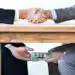 underthetable-transactions-cropped-shot-two-businessmen-shaking-hands-while-money-passes-hands-table - Pratama Institute