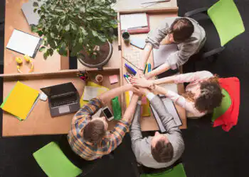 Group of five colleagues around a table joining hands in a teamwork gesture, viewed from above.