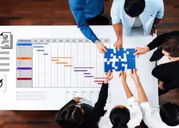 Overhead view of a team collaborating over a project Gantt chart, placing a blue puzzle piece on the timeline on a white table.