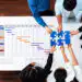 Overhead view of a team collaborating over a project Gantt chart, placing a blue puzzle piece on the timeline on a white table.