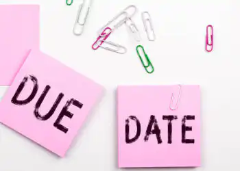 Two pink sticky notes spell 'DUE' and 'DATE' on a white desk with colorful paperclips scattered around.