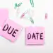 Two pink sticky notes spell 'DUE' and 'DATE' on a white desk with colorful paperclips scattered around.