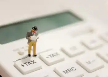 Miniature businessman stands on a calculator key labeled +TAX, looking at papers to symbolize tax calculation.