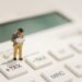 Miniature businessman stands on a calculator key labeled +TAX, looking at papers to symbolize tax calculation.