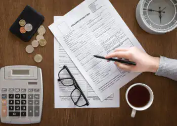 Hand writing on visa application forms on a wooden desk, with a calculator, glasses, coins, wallet, clock, and coffee nearby.