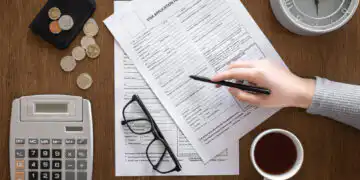 Hand writing on visa application forms on a wooden desk, with a calculator, glasses, coins, wallet, clock, and coffee nearby.