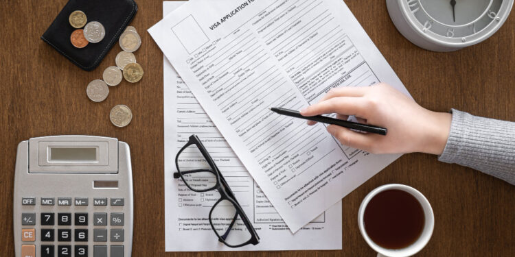 Hand writing on visa application forms on a wooden desk, with a calculator, glasses, coins, wallet, clock, and coffee nearby.