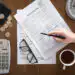 Ilustrasi penerimaan pajak - Pratama Institute Hand writing on visa application forms on a wooden desk, with a calculator, glasses, coins, wallet, clock, and coffee nearby.