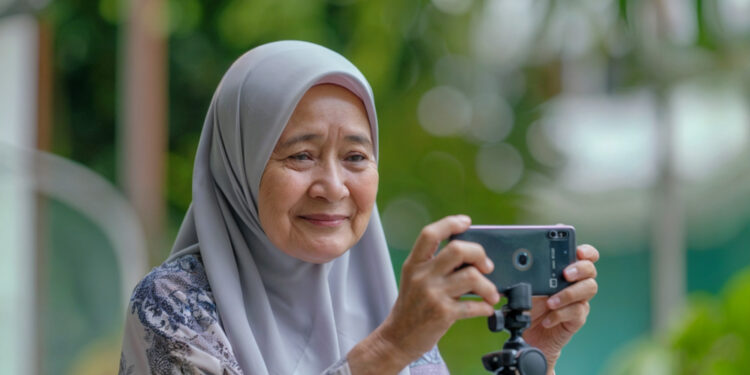 Older woman in a light gray hijab and patterned blouse smiling as she uses a smartphone mounted on a small tripod outdoors.”,