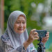 Older woman in a light gray hijab and patterned blouse smiling as she uses a smartphone mounted on a small tripod outdoors.”,