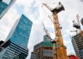 - Pratama Institute Tall yellow construction cranes loom over partially built high-rise buildings in a cityscape against a partly cloudy sky, modern glass towers nearby.