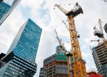 Tall yellow construction cranes loom over partially built high-rise buildings in a cityscape against a partly cloudy sky, modern glass towers nearby.