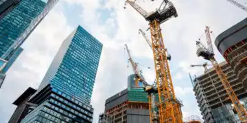 Tall yellow construction cranes loom over partially built high-rise buildings in a cityscape against a partly cloudy sky, modern glass towers nearby.