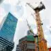 Tall yellow construction cranes loom over partially built high-rise buildings in a cityscape against a partly cloudy sky, modern glass towers nearby.