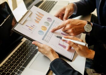 Business professional pointing to colorful charts on a clipboard beside a laptop, reviewing data.