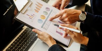 Business professional pointing to colorful charts on a clipboard beside a laptop, reviewing data.