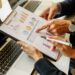 - Pratama Institute Business professional pointing to colorful charts on a clipboard beside a laptop, reviewing data.