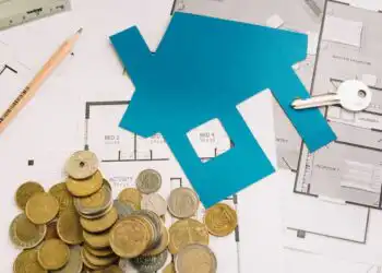 Blue house cutout on floor plans with coins, a key, pencil, and ruler—evoking home financing or real estate budgeting.