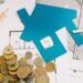 Blue house cutout on floor plans with coins, a key, pencil, and ruler—evoking home financing or real estate budgeting.