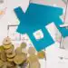 - Pratama Institute Blue house cutout on floor plans with coins, a key, pencil, and ruler—evoking home financing or real estate budgeting.
