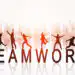 Silhouetted people jumping on the letters spelling TEAMWORK against a city skyline at sunrise, with a reflective surface below.
