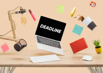 Laptop suspended above a cluttered desk with 'DEADLINE' on the screen, surrounded by sticky notes, a mug, lamp, plant, and notebook.