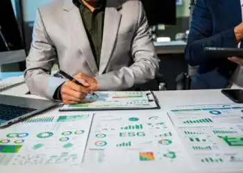 Two professionals review ESG reports and charts spread across a conference table, one writing with a pen while the other holds a tablet/light device nearby.