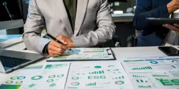 Two professionals review ESG reports and charts spread across a conference table, one writing with a pen while the other holds a tablet/light device nearby.