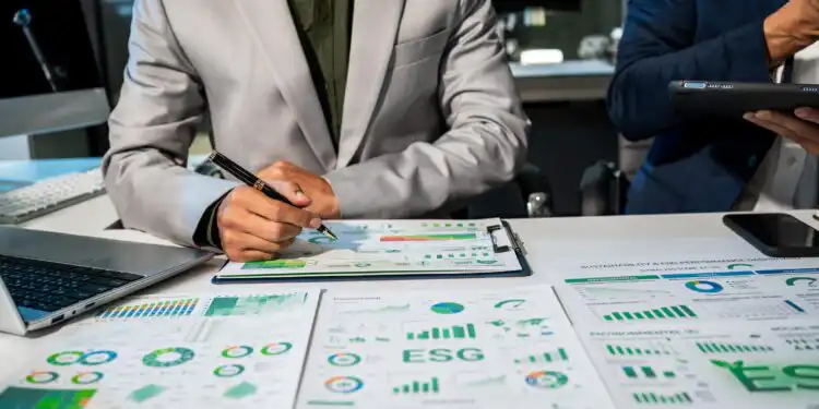 Two professionals review ESG reports and charts spread across a conference table, one writing with a pen while the other holds a tablet/light device nearby.