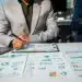 - Pratama Institute Two professionals review ESG reports and charts spread across a conference table, one writing with a pen while the other holds a tablet/light device nearby.