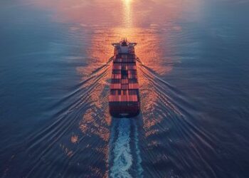 Container ship sailing at sunset on calm water, with a plane flying overhead and a distant bridge.