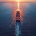 Container ship sailing at sunset on calm water, with a plane flying overhead and a distant bridge.