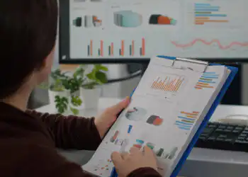 Person reviews printed charts on a clipboard while a computer monitor shows data dashboards in an office setting.