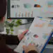 Person reviews printed charts on a clipboard while a computer monitor shows data dashboards in an office setting.
